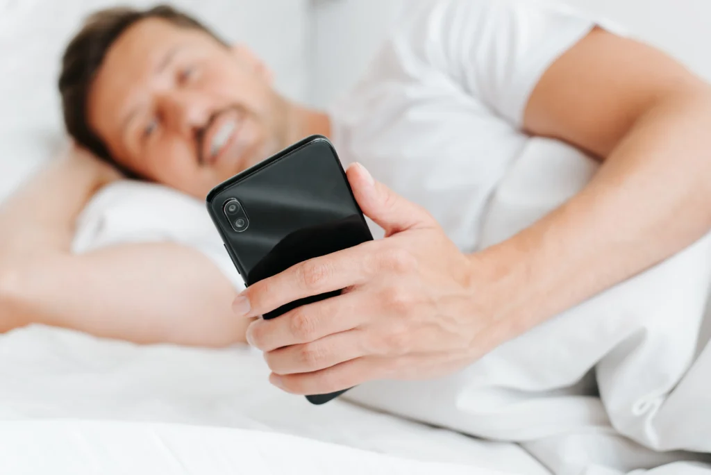 Hombre recostado en la cama sosteniendo su smartphone con una sonrisa